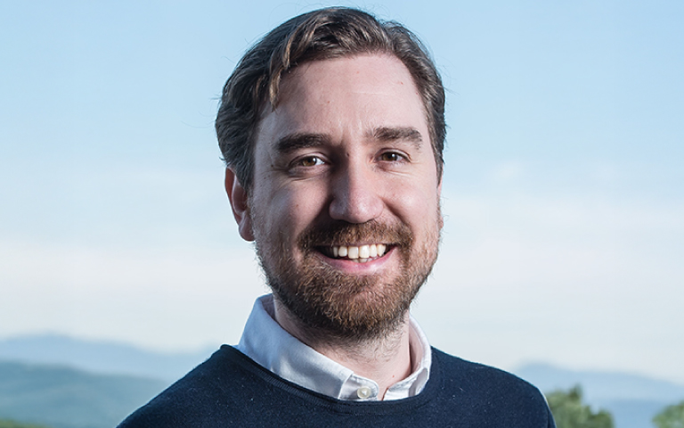 Jonathan Graves, a smiling man with short brown hair and a beard, wearing a white shirt under a navy sweater, stands outdoors with a blue sky and hills in the background.