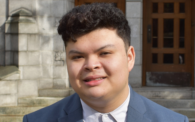 Kareem Hassib, a young man with short dark wavy hair, wears a navy blue suit jacket and white shirt while standing outside a stone building.